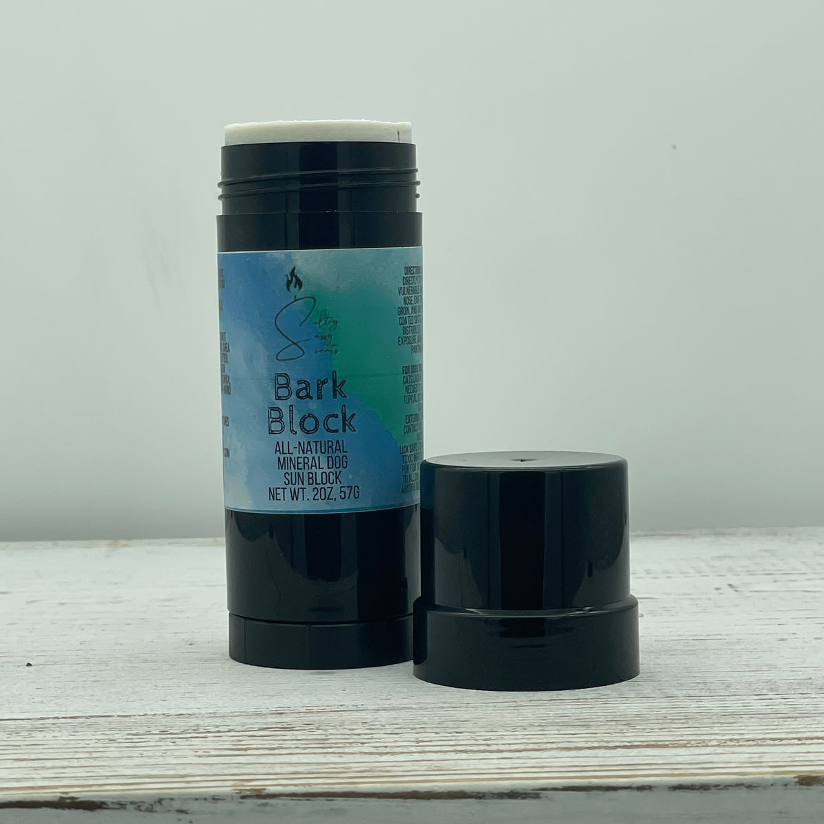 An open 2-ounce black twist-up stick container of Sultry Sassy Scents Bark Block All-Natural Mineral Dog Sun Block, showing the white balm product next to its black plastic cap on a light wooden table.