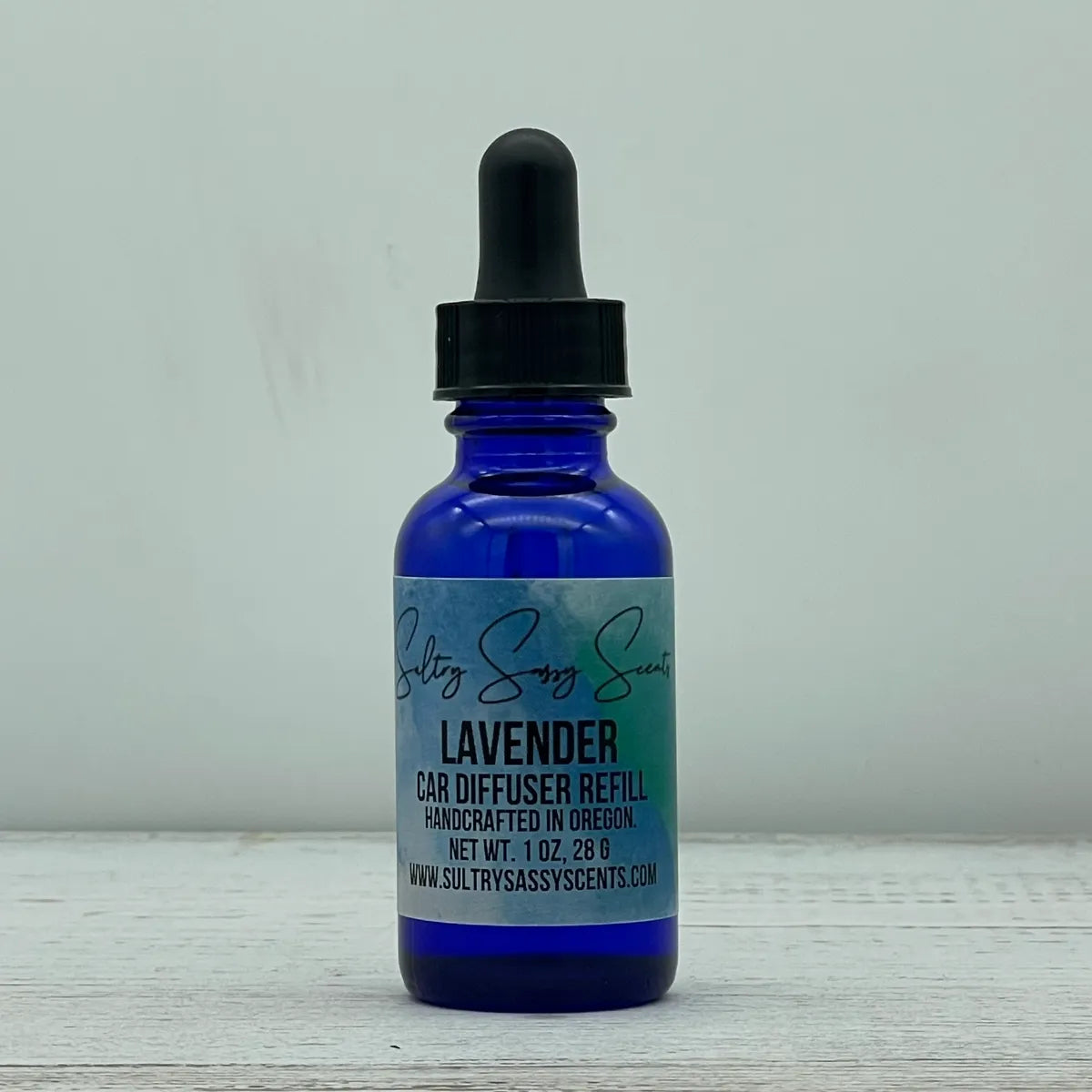 Front label view of a cobalt blue glass dropper bottle containing Sultry Sassy Scents "Lavender" Car Diffuser Refill, centered on a light wooden background.