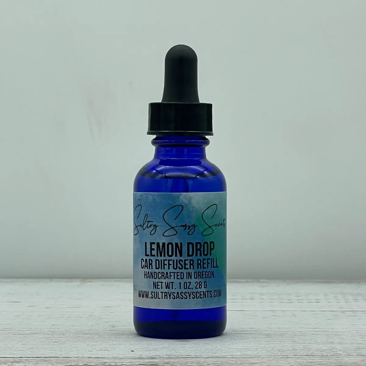 Front label view of a cobalt blue glass dropper bottle containing Sultry Sassy Scents "Lemon Drop" Car Diffuser Refill, highlighting the bold product name layout on a white wood surface.