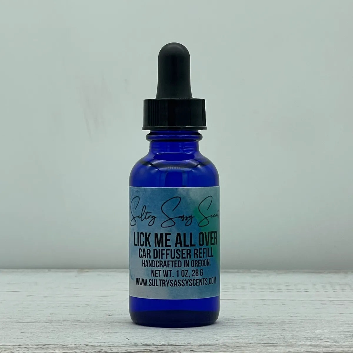 Front label view of a cobalt blue glass dropper bottle containing Sultry Sassy Scents "Lick Me All Over" Car Diffuser Refill, sitting on a light wood grain surface.