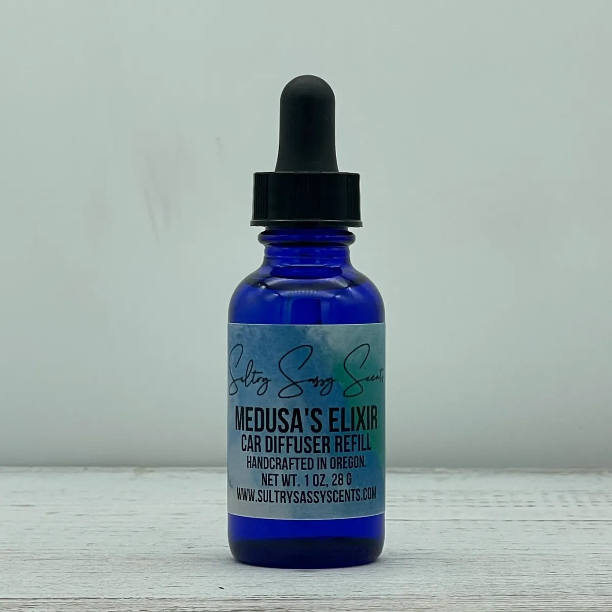Front label view of a cobalt blue glass dropper bottle containing Sultry Sassy Scents "Medusa's Elixir" Car Diffuser Refill, centered against a light gray backdrop.