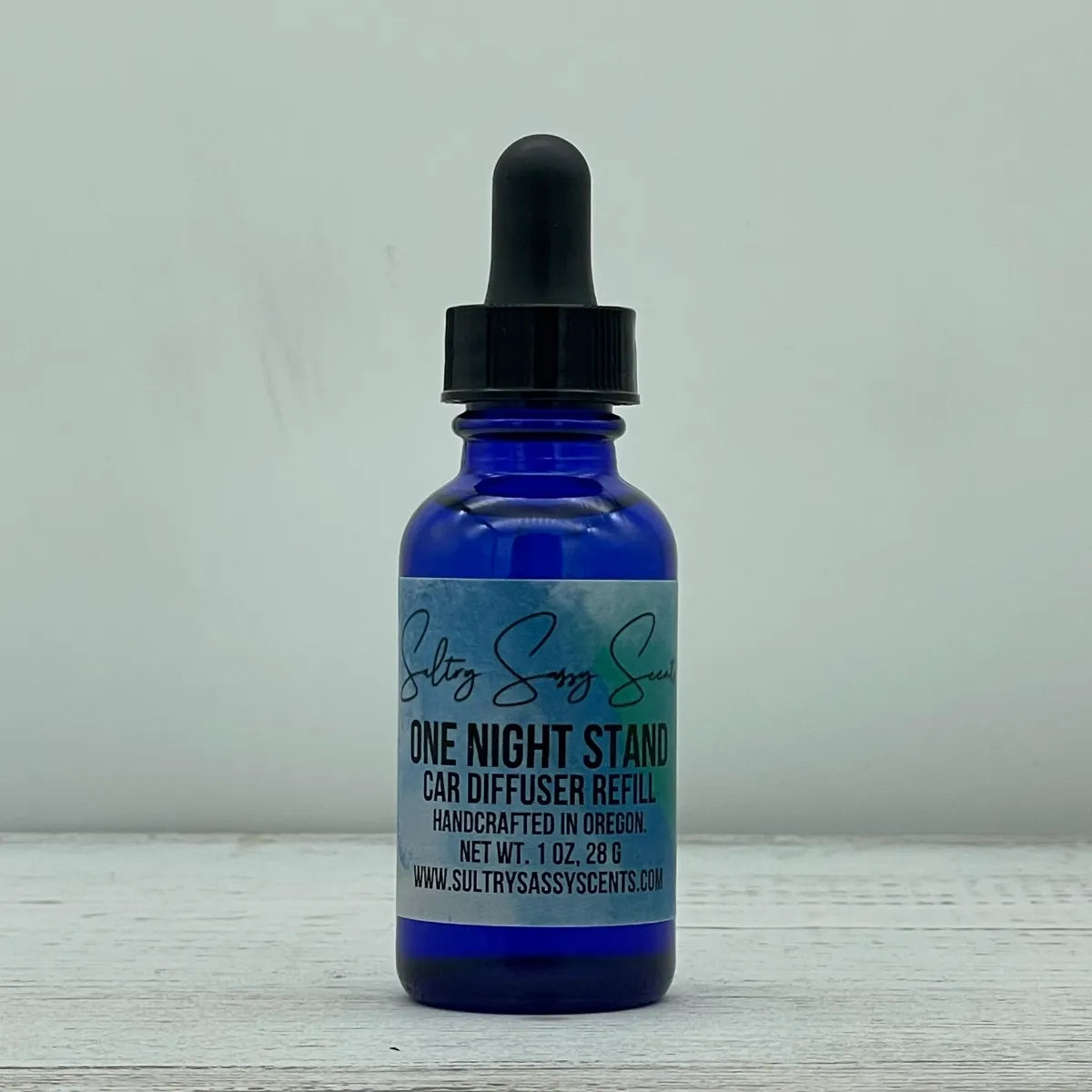 Front label view of a cobalt blue glass dropper bottle containing Sultry Sassy Scents "One Night Stand" Car Diffuser Refill, positioned on a rustic light wood table.
