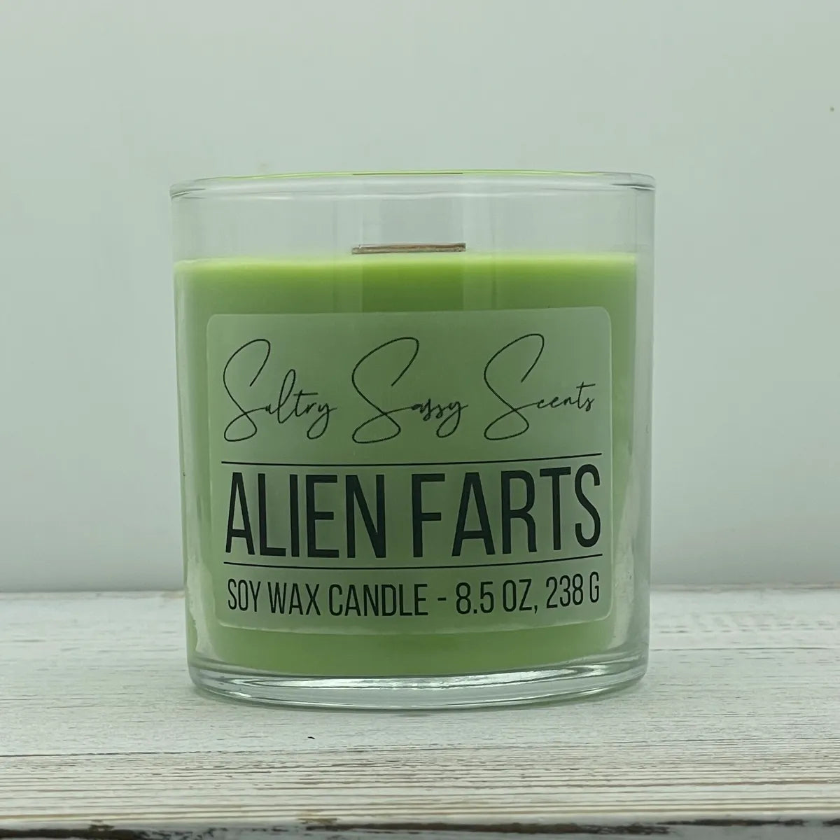 An 8.5-ounce jar of Sultry Sassy Scents "Alien Farts" soy wax candle featuring a bright green wax color and a wooden wick, centered on a white wood surface.