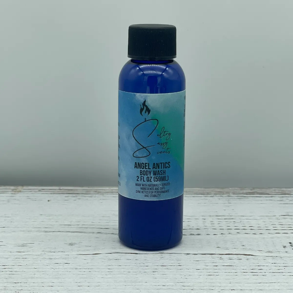 Front label view of a small 2-ounce travel size blue bottle with a black screw cap containing Sultry Sassy Scents "Angel Antics" Body Wash, centered on a light wood backdrop.