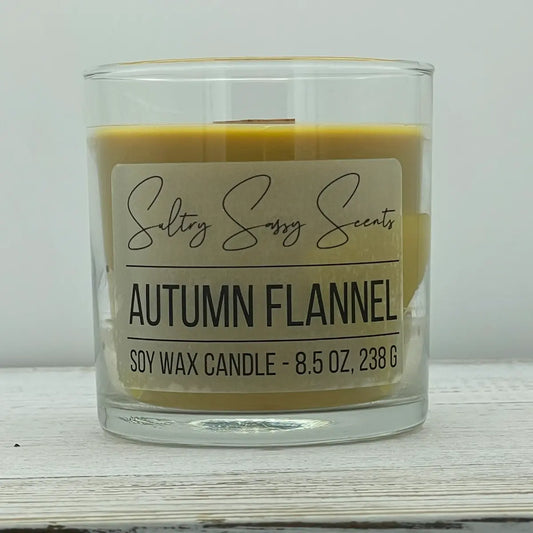 An 8.5-ounce clear glass jar of Sultry Sassy Scents "Autumn Flannel" soy wax candle featuring a smooth mustard-yellow wax color and a flat wooden wick, centered on a white wood background.