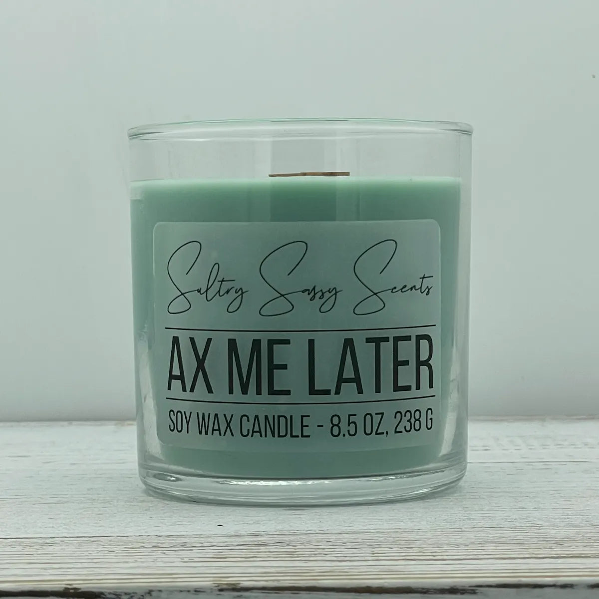 An 8.5-ounce jar of Sultry Sassy Scents "Ax Me Later" soy wax candle featuring a pale sage green wax color and a wooden wick, resting on a white-washed wood table.