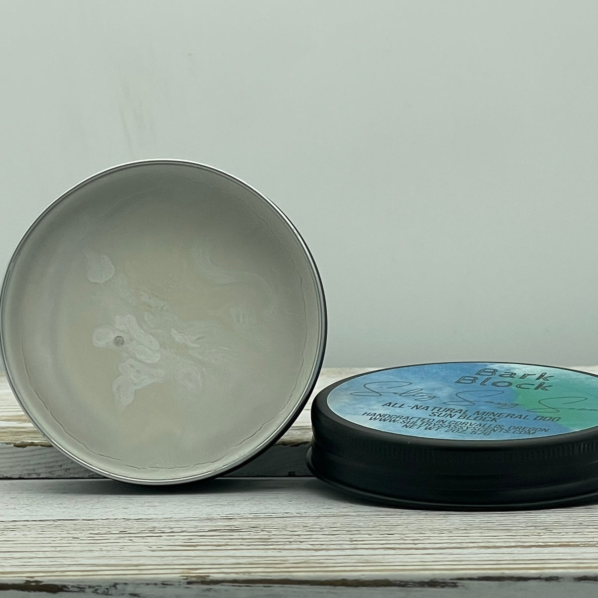 An open 2-ounce round metal tin of Sultry Sassy Scents Bark Block All-Natural Mineral Dog Sun Block, showing the smooth white mineral sunblock cream inside next to its decorated black lid.
