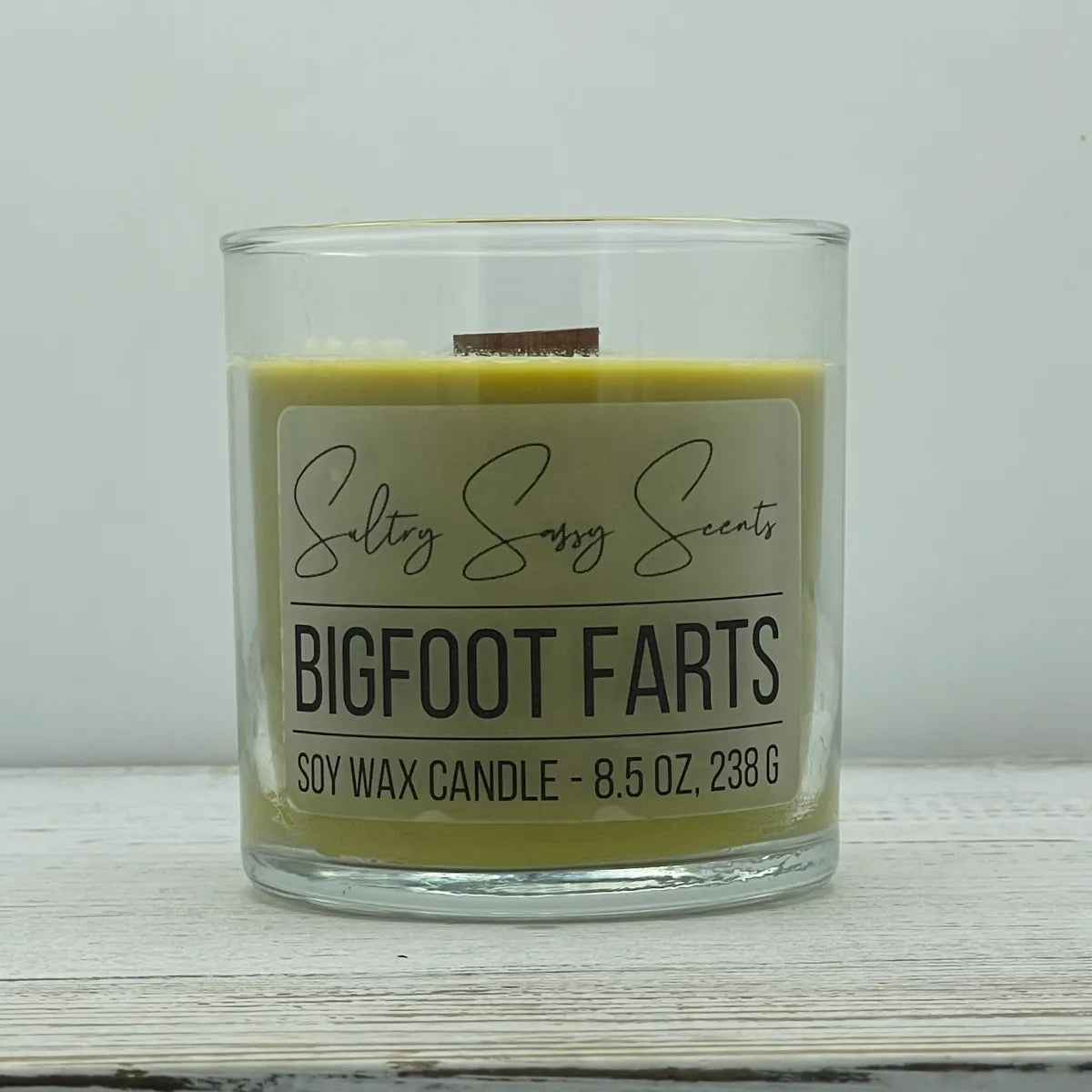 An 8.5-ounce jar of Sultry Sassy Scents "Bigfoot Farts" soy wax candle featuring an olive green wax color and a wooden wick, resting on a white wood surface.