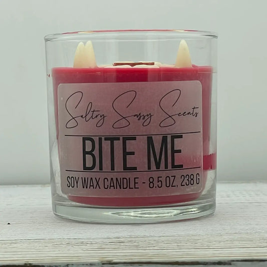 An 8.5-ounce clear glass jar of Sultry Sassy Scents "Bite Me" soy wax candle featuring crimson red wax decorated with two pointed white vampire fang wax toppers embedded on top.