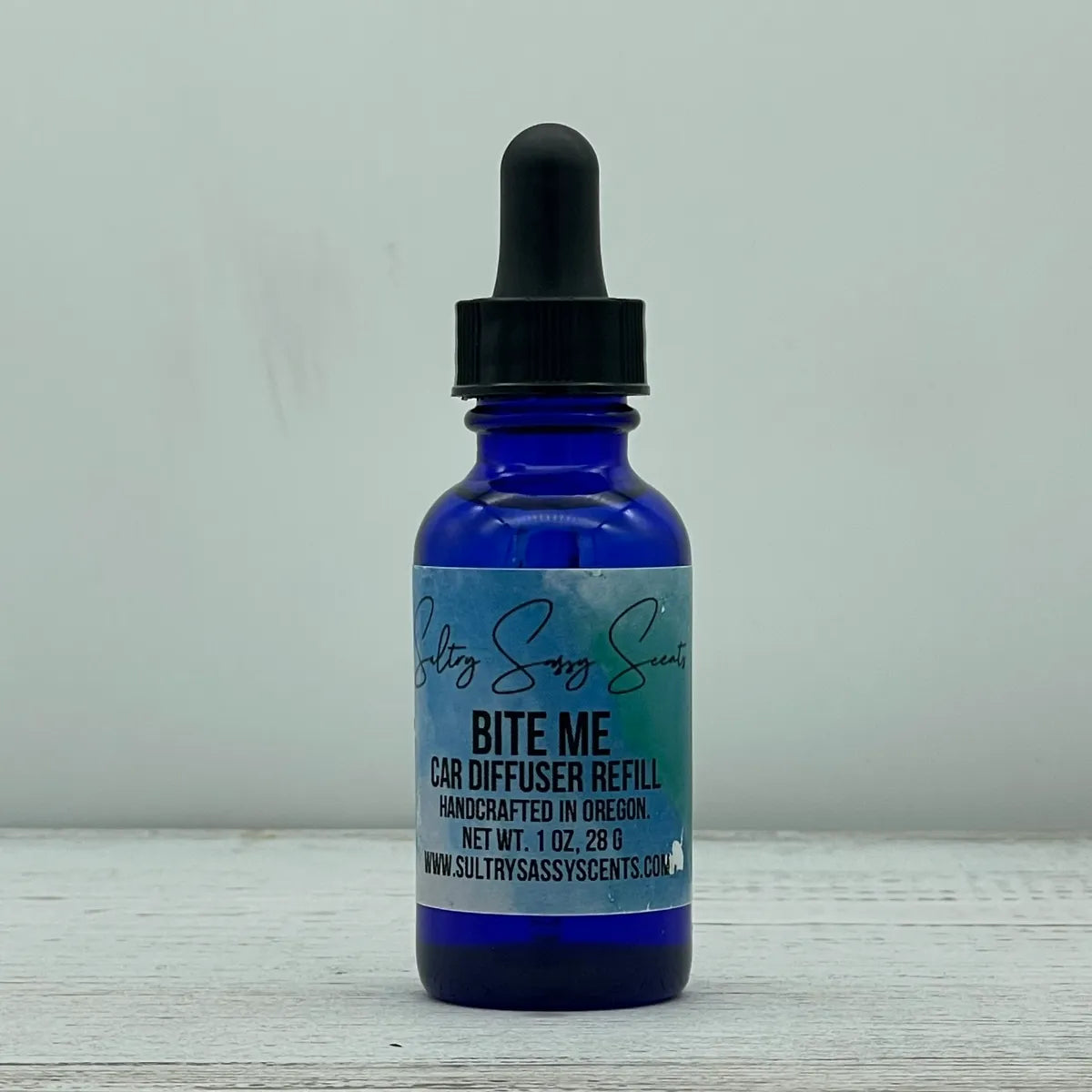 Front label view of a blue glass dropper bottle for Sultry Sassy Scents "Bite Me" Car Diffuser Refill, displayed on a light wooden background.