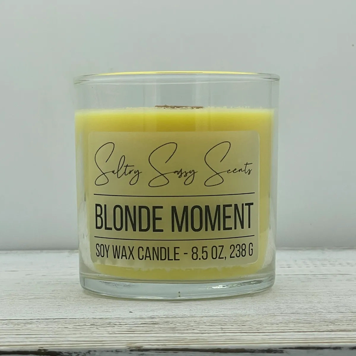 An 8.5-ounce jar of Sultry Sassy Scents "Blonde Moment" soy wax candle featuring a soft pastel yellow wax color and a wooden wick, displayed on a rustic light wood table.