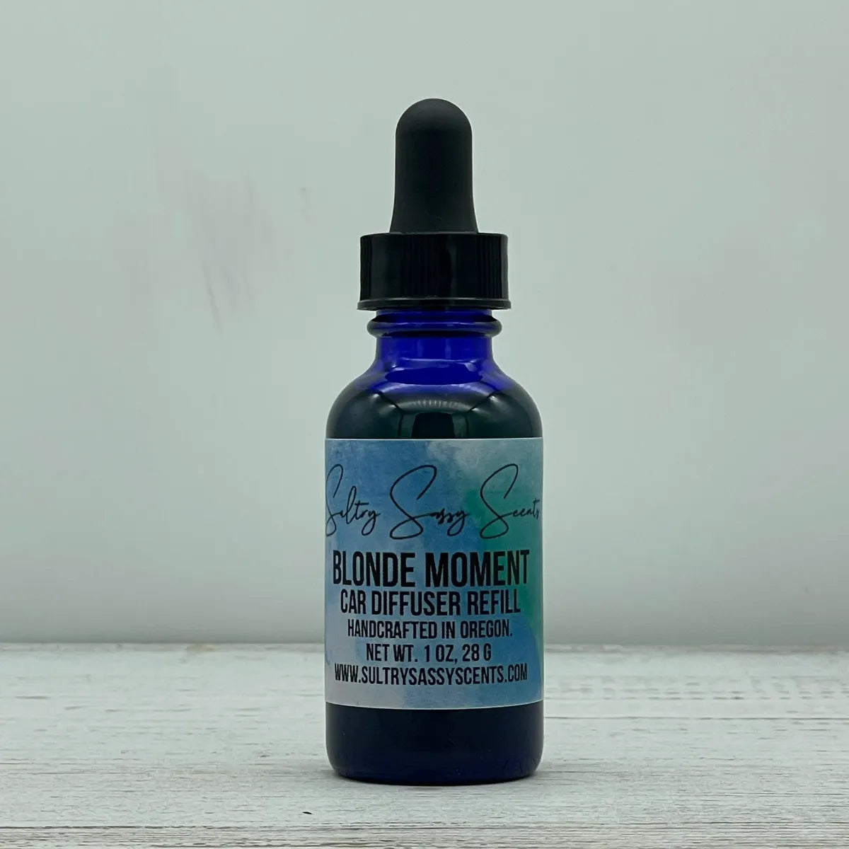 Front label view of a blue glass dropper bottle for Sultry Sassy Scents "Blonde Moment" Car Diffuser Refill, centered on a rustic white wood table.
