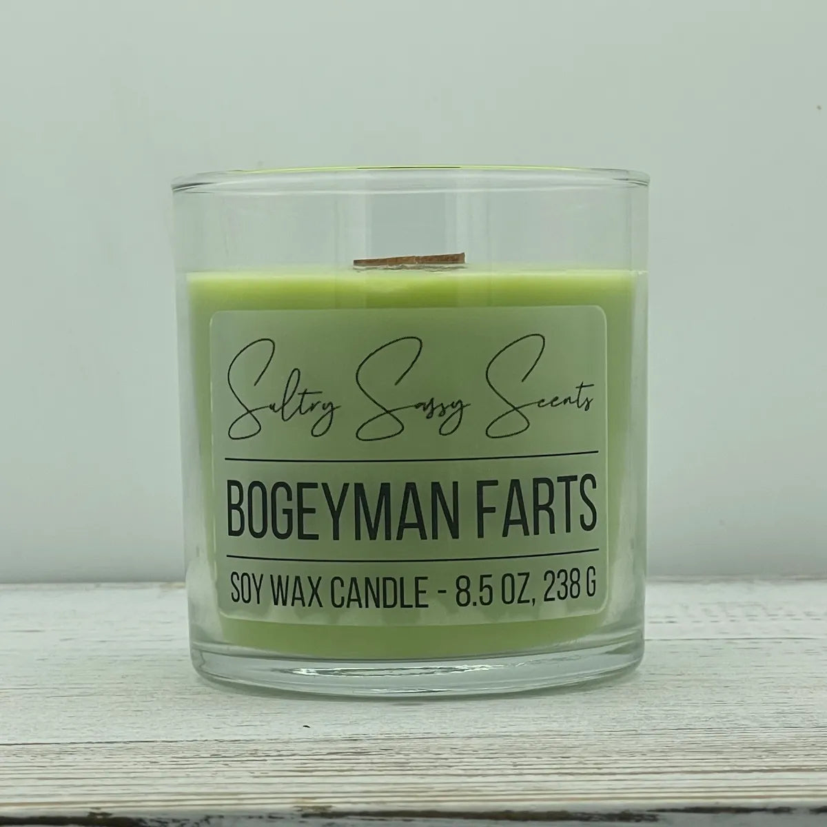 An 8.5-ounce jar of Sultry Sassy Scents "Bogeyman Farts" soy wax candle featuring a light mint green wax color and a wooden wick, centered on a white wood background.