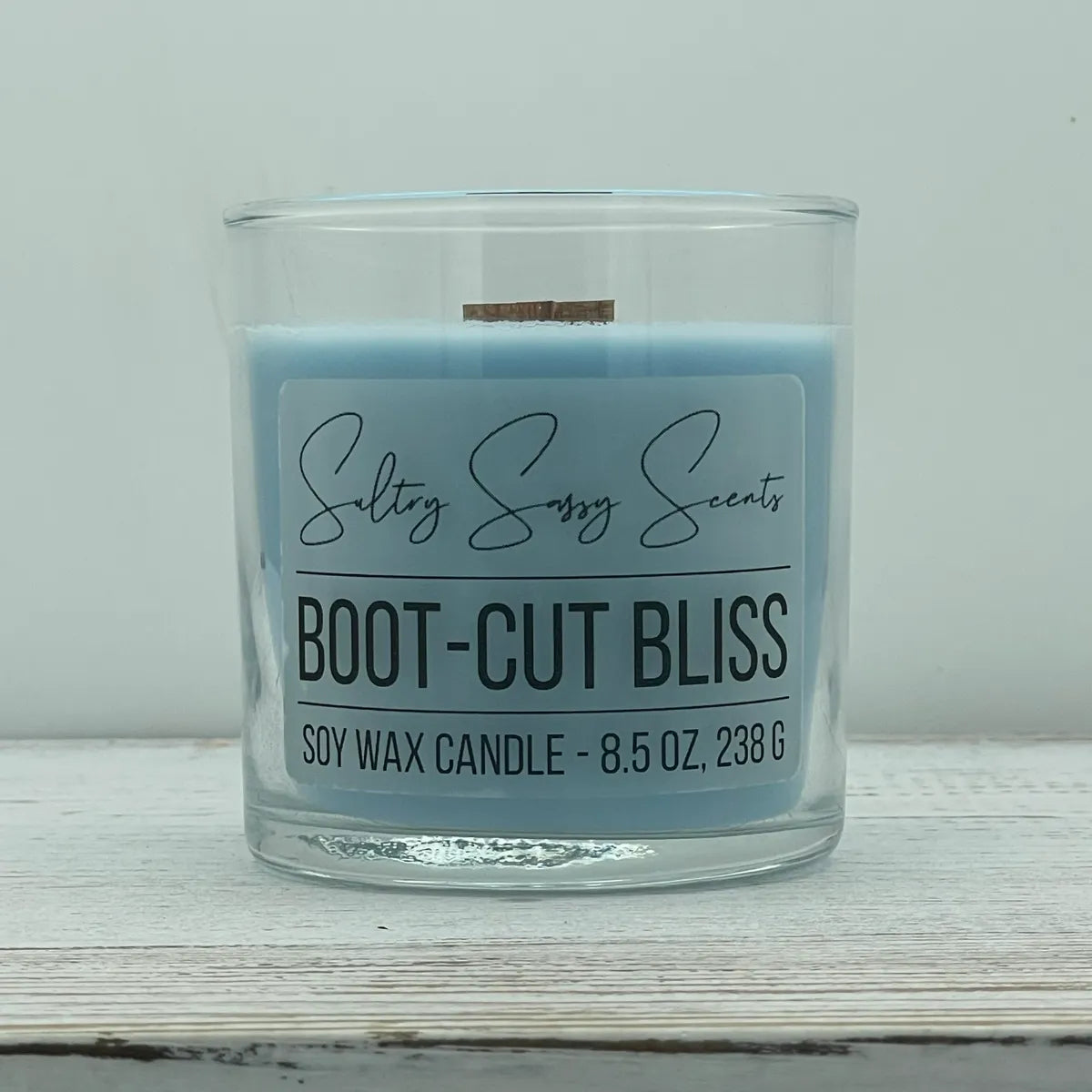 An 8.5-ounce clear glass jar of Sultry Sassy Scents "Boot-Cut Bliss" soy wax candle featuring a light denim blue wax color and a flat wooden wick, centered on a white wooden table.