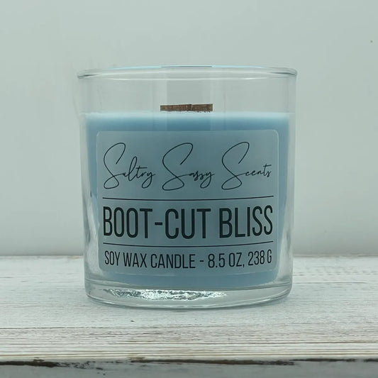 An 8.5-ounce clear glass jar of Sultry Sassy Scents "Boot-Cut Bliss" soy wax candle featuring a light denim blue wax color and a flat wooden wick, centered on a white wooden table.