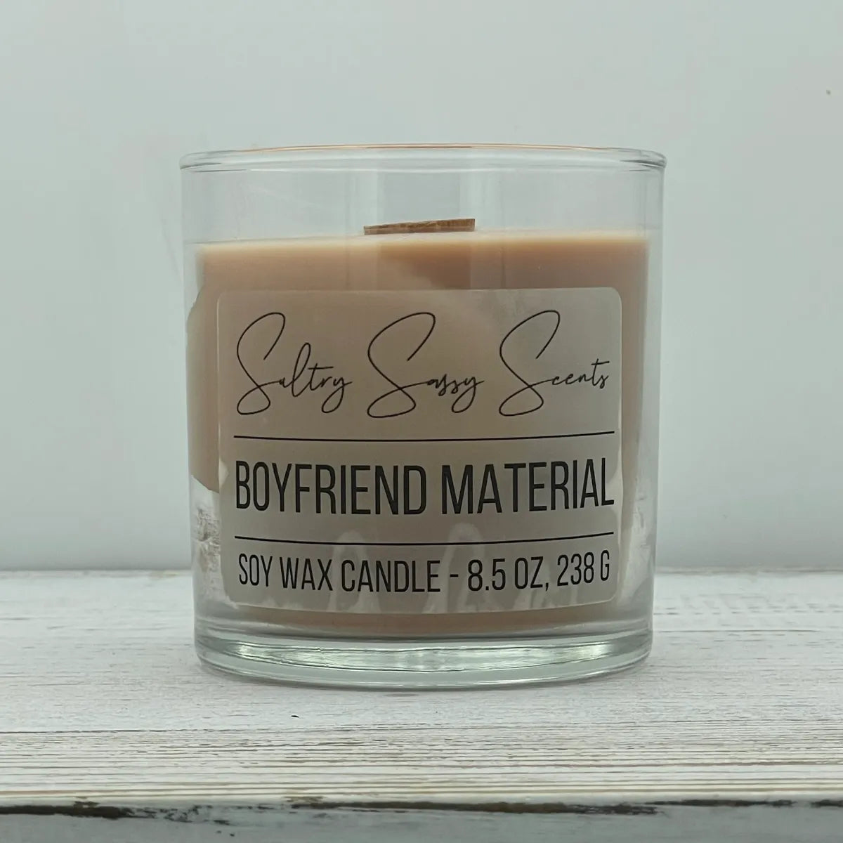 An 8.5-ounce clear glass jar of Sultry Sassy Scents "Boyfriend Material" soy wax candle showcasing a smooth, light tan wax color with a wooden wick, displayed on a rustic wood background.