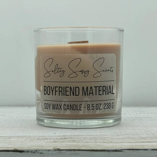 An 8.5-ounce clear glass jar of Sultry Sassy Scents "Boyfriend Material" soy wax candle showcasing a smooth, light tan wax color with a wooden wick, displayed on a rustic wood background.