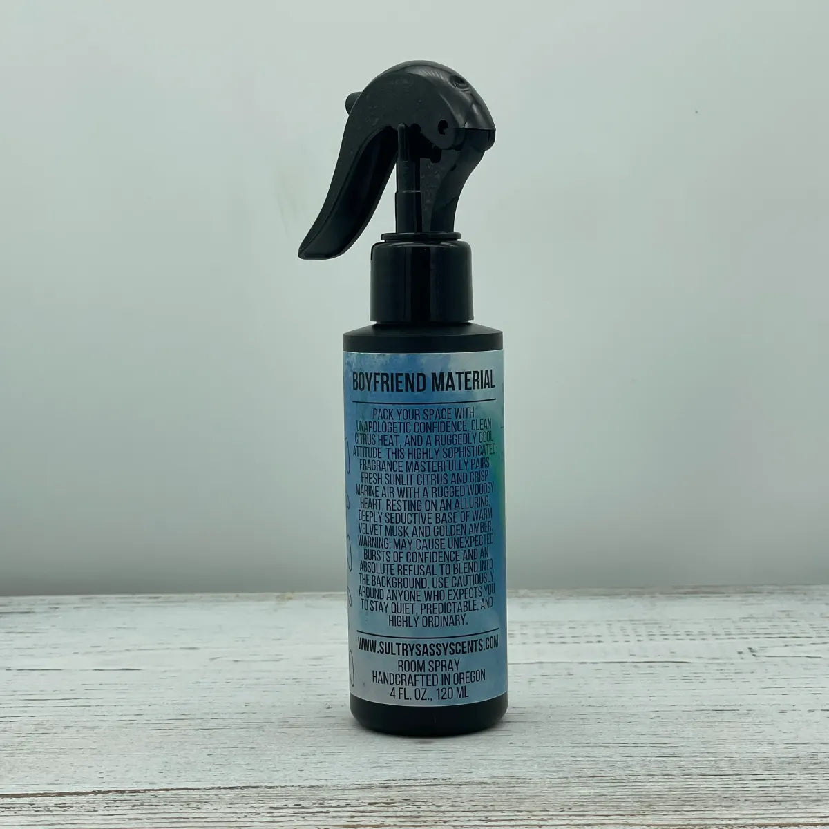 Front label of a black spray bottle for Sultry Sassy Scents "Boyfriend Material" Room Spray, showcasing bold lettering and a clean paragraph block detailing a fresh citrus and woodsy musk fragrance.