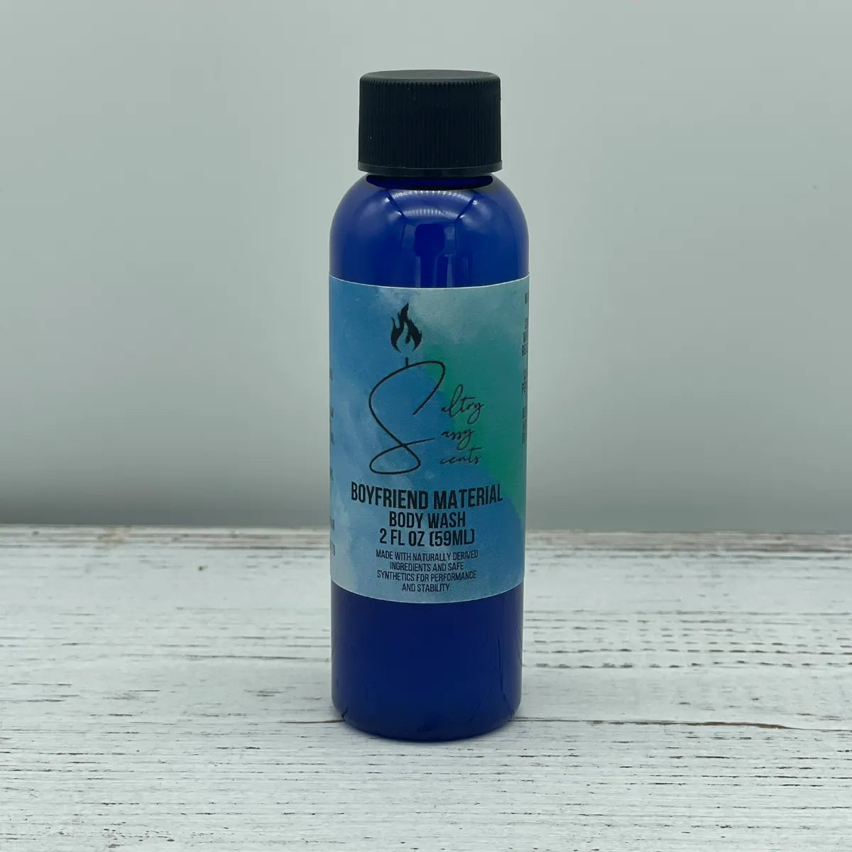 Front label view of a small 2-ounce travel size blue bottle with a black screw cap containing Sultry Sassy Scents "Boyfriend Material" Body Wash, resting on a white wood surface.