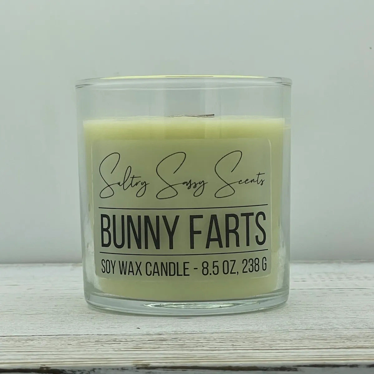 An 8.5-ounce clear glass jar of Sultry Sassy Scents "Bunny Farts" soy wax candle displaying a soft pastel yellow-green wax color and a flat wooden wick, resting on a white-washed wood table.