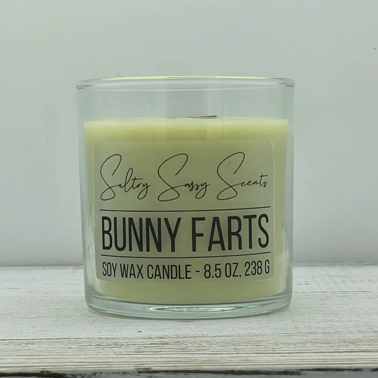 An 8.5-ounce clear glass jar of Sultry Sassy Scents "Bunny Farts" soy wax candle displaying a soft pastel yellow-green wax color and a flat wooden wick, resting on a white-washed wood table.