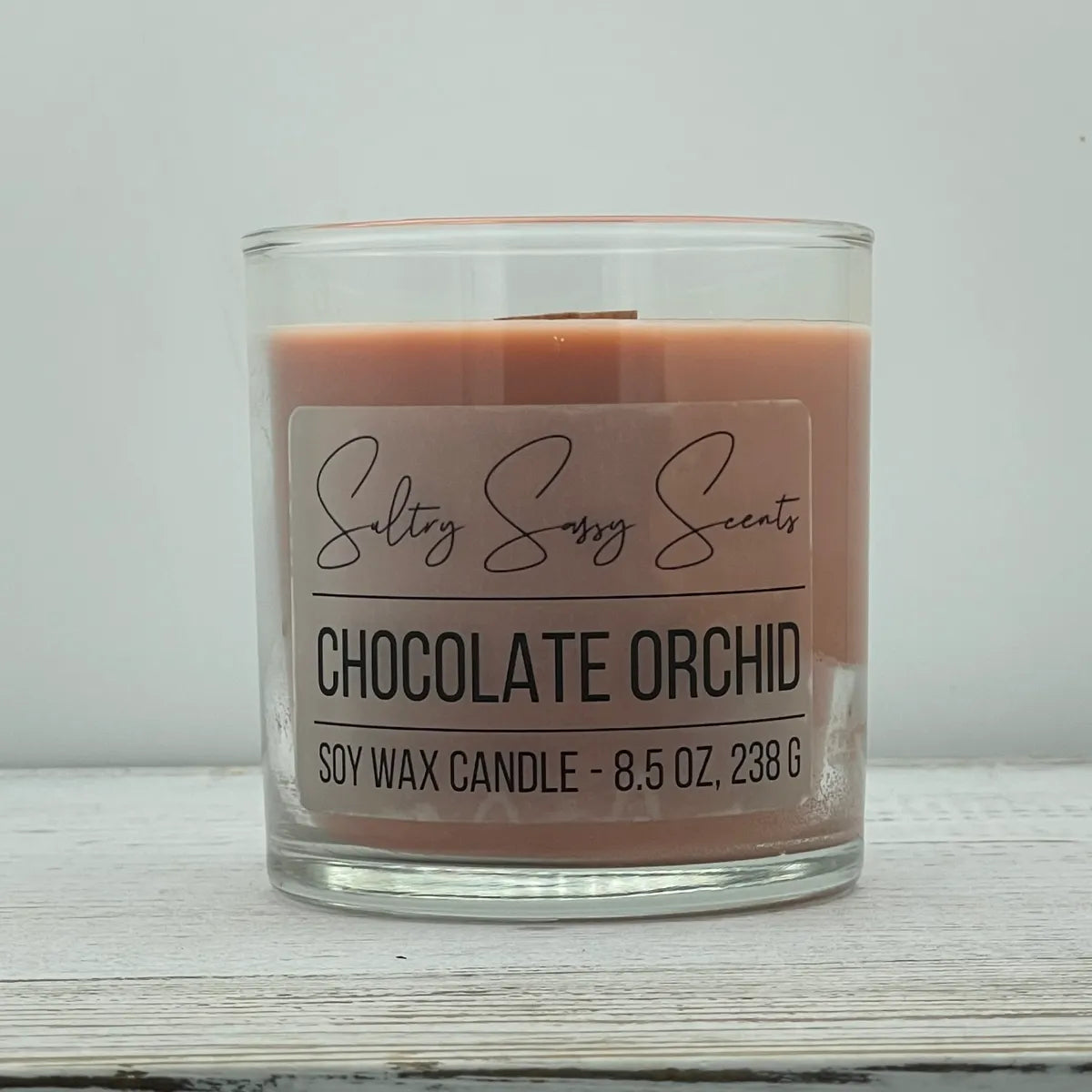 An 8.5-ounce clear glass jar of Sultry Sassy Scents "Chocolate Orchid" soy wax candle showcasing a smooth warm taupe wax color with a flat wooden wick, displayed on a rustic wood surface.