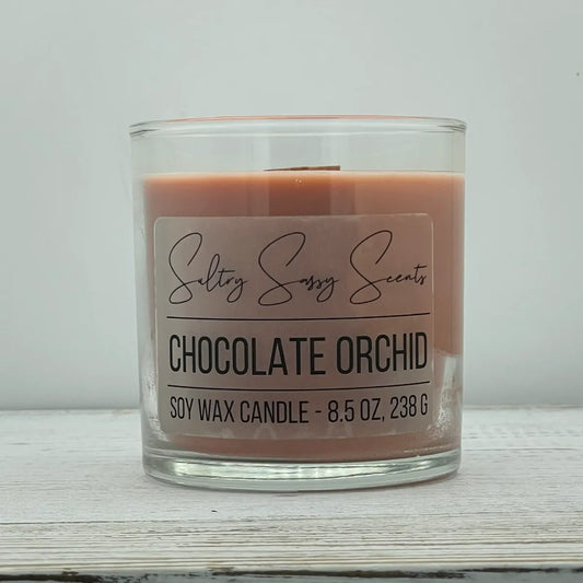 An 8.5-ounce clear glass jar of Sultry Sassy Scents "Chocolate Orchid" soy wax candle showcasing a smooth warm taupe wax color with a flat wooden wick, displayed on a rustic wood surface.