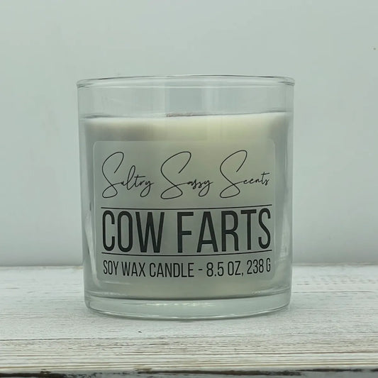 An 8.5-ounce clear glass jar of Sultry Sassy Scents "Cow Farts" soy wax candle featuring a soft off-white wax color and a flat wooden wick, resting on a white wooden surface.