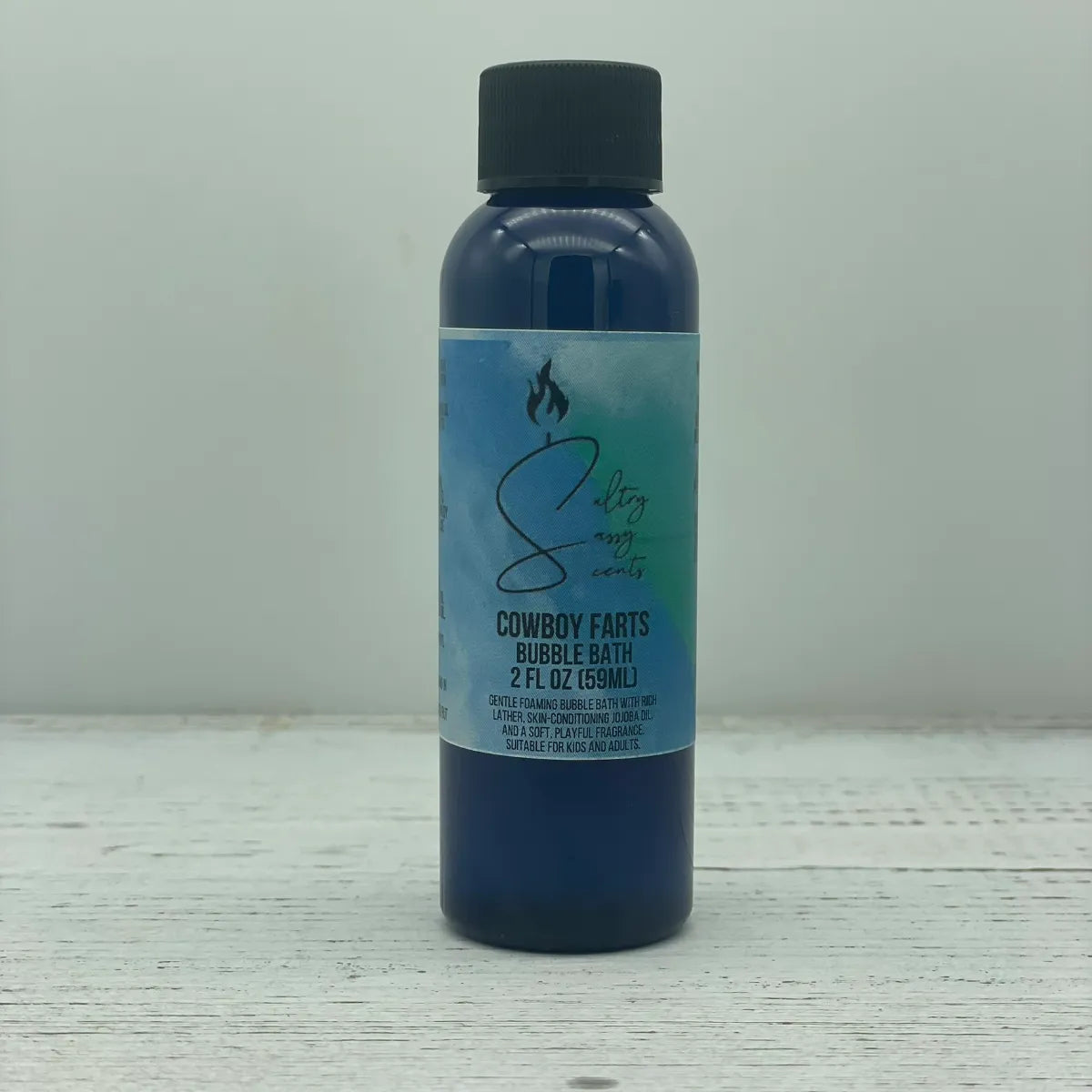 Front label view of a small 2-ounce travel size blue bottle with a black screw cap containing Sultry Sassy Scents "Cowboy Farts" Bubble Bath, centered on a rustic wood surface.