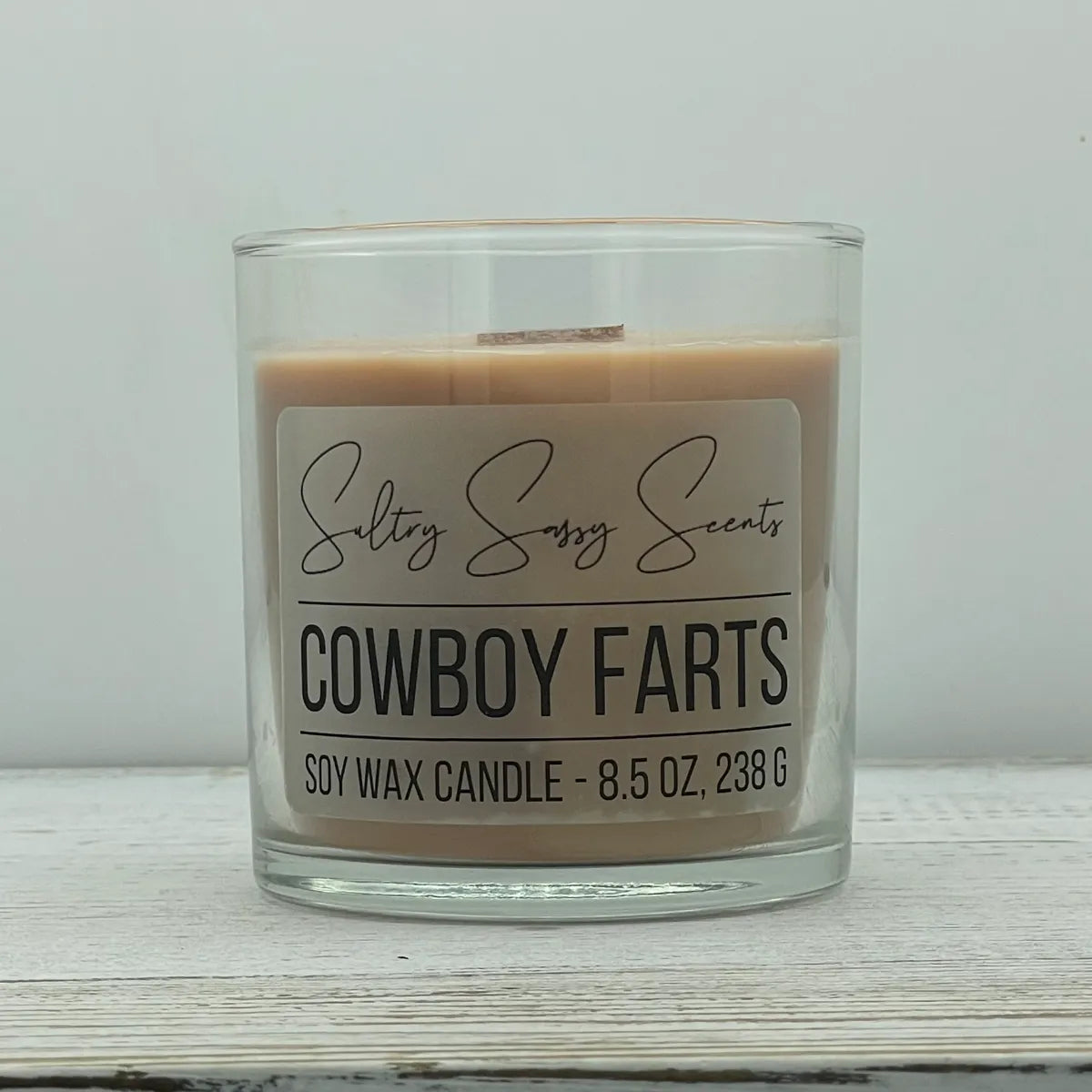 An 8.5-ounce clear glass jar of Sultry Sassy Scents "Cowboy Farts" soy wax candle displaying a smooth light tan wax color, centered on a light wooden background.