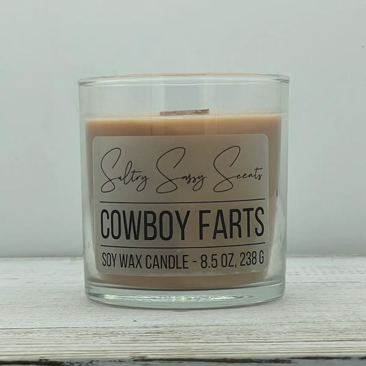 An 8.5-ounce clear glass jar of Sultry Sassy Scents "Cowboy Farts" soy wax candle displaying a smooth light tan wax color, centered on a light wooden background.