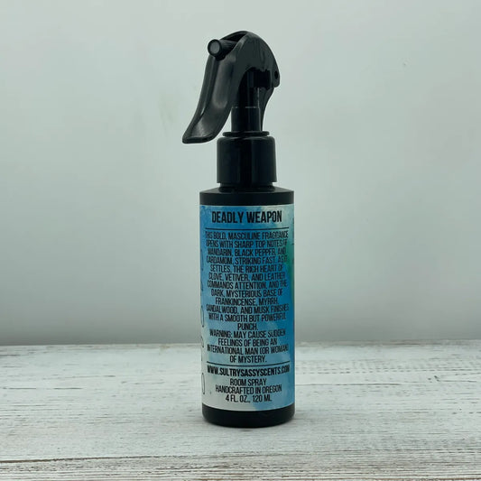 Front label of a black spray bottle for Sultry Sassy Scents "Deadly Weapon" Room Spray, featuring stylized text describing a bold, masculine fragrance with mandarin, black pepper, clove, leather, and frankincense.