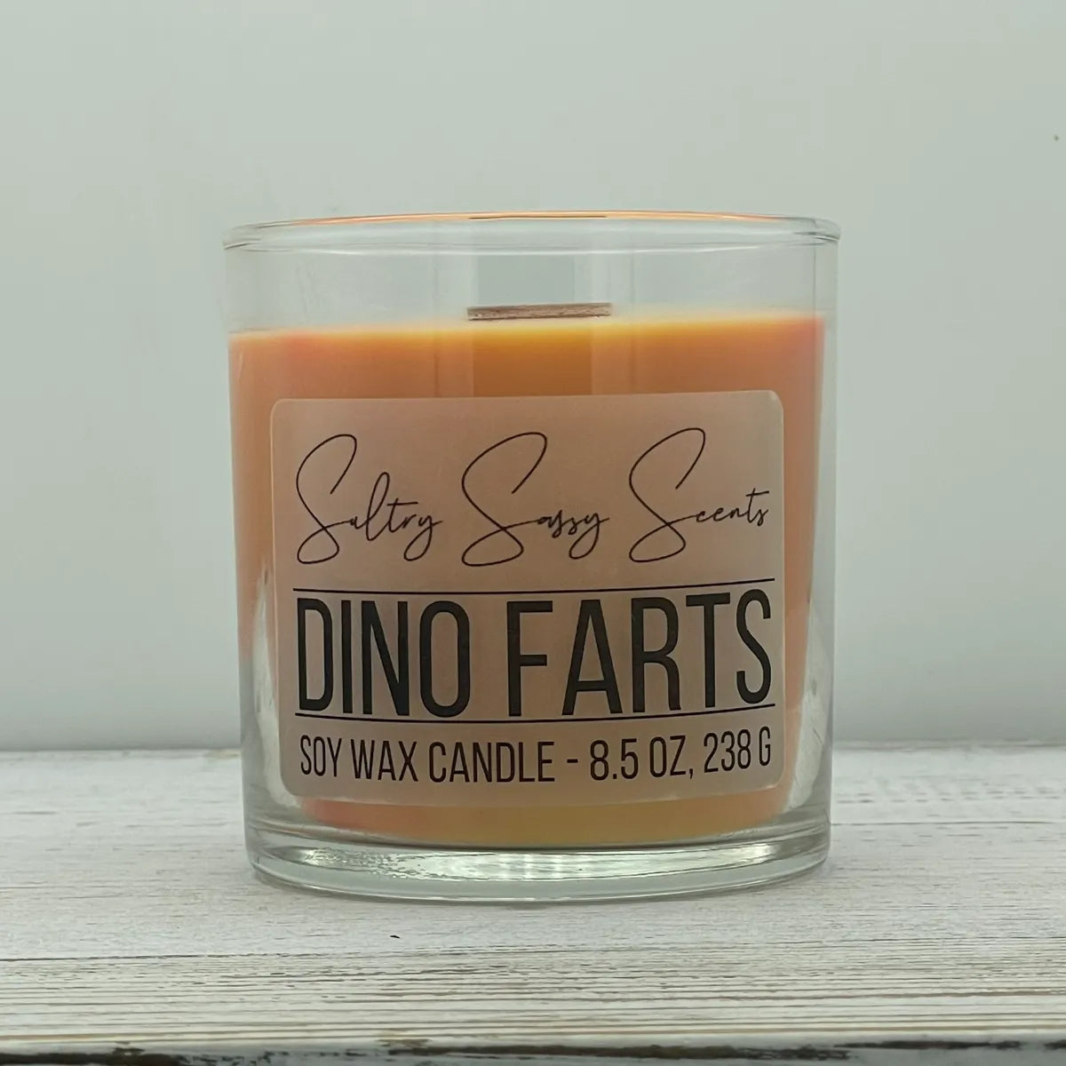 An 8.5-ounce clear glass jar of Sultry Sassy Scents "Dino Farts" soy wax candle showcasing a warm terracotta orange wax color and a flat wooden wick.