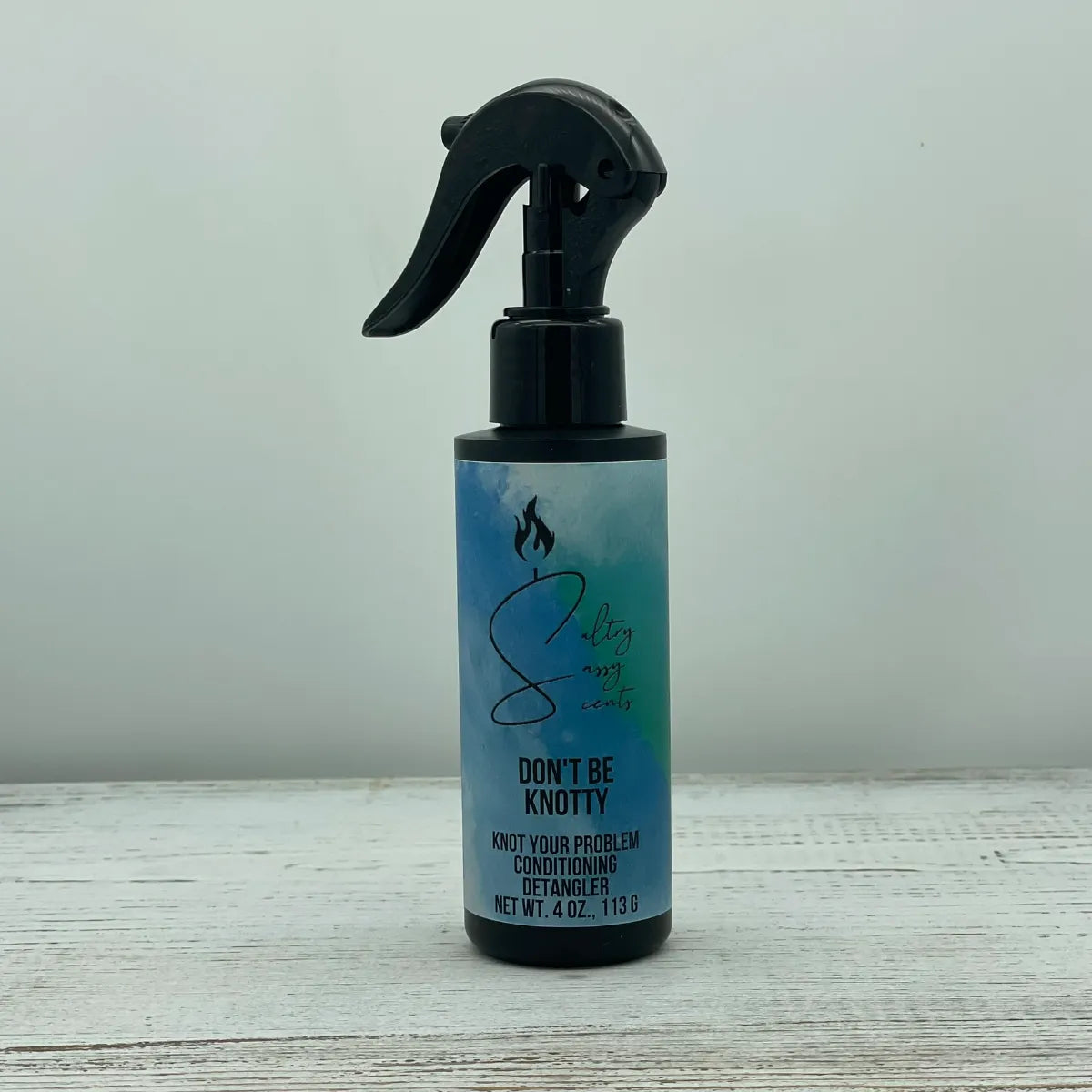 A single black spray bottle of Sultry Sassy Scents Knot Your Problem Conditioning Detangler in the Don't Be Knotty variation, centered on a light wood background.