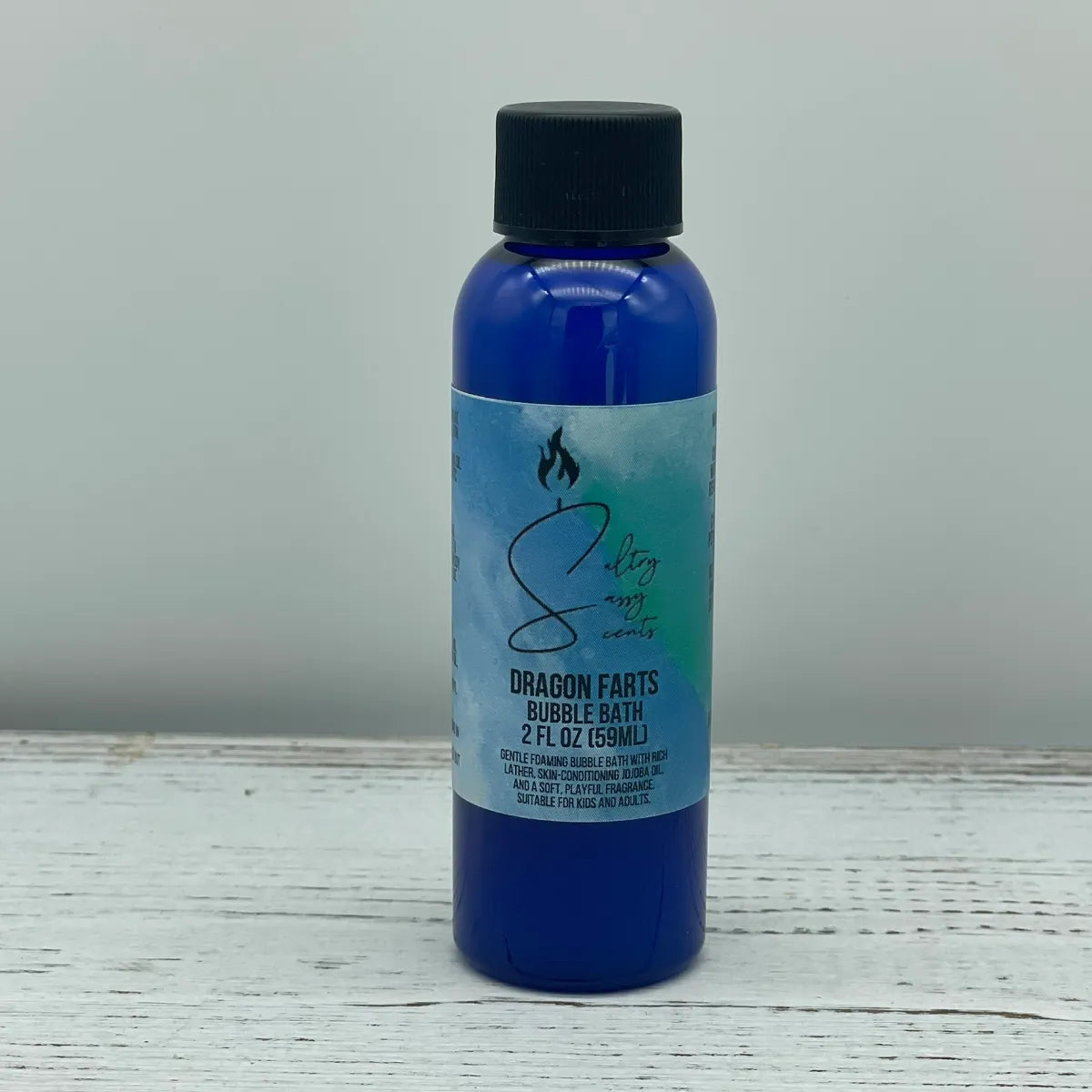 Front label view of a small 2-ounce travel size blue bottle with a black screw cap containing Sultry Sassy Scents "Dragon Farts" Bubble Bath, displayed on a light wooden background.