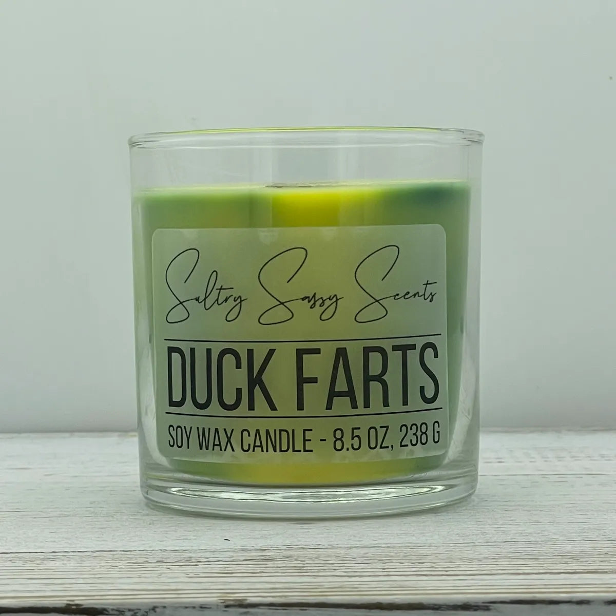 An 8.5-ounce clear glass jar of Sultry Sassy Scents "Duck Farts" soy wax candle showcasing a unique, multi-toned swirl of green and bright yellow wax with a wooden wick.