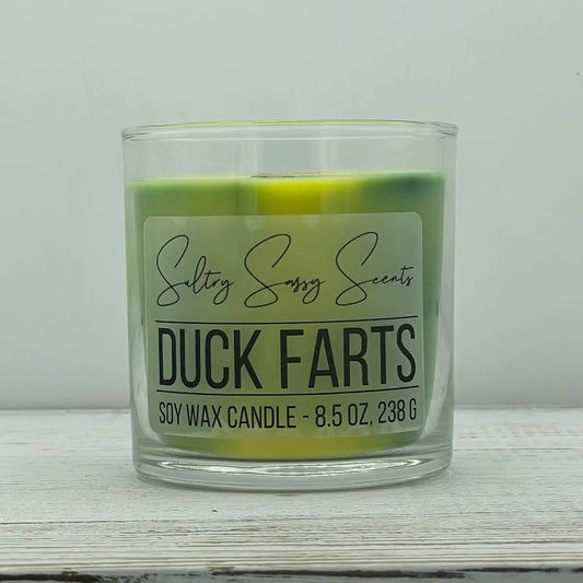 An 8.5-ounce clear glass jar of Sultry Sassy Scents "Duck Farts" soy wax candle showcasing a unique, multi-toned swirl of green and bright yellow wax with a wooden wick.