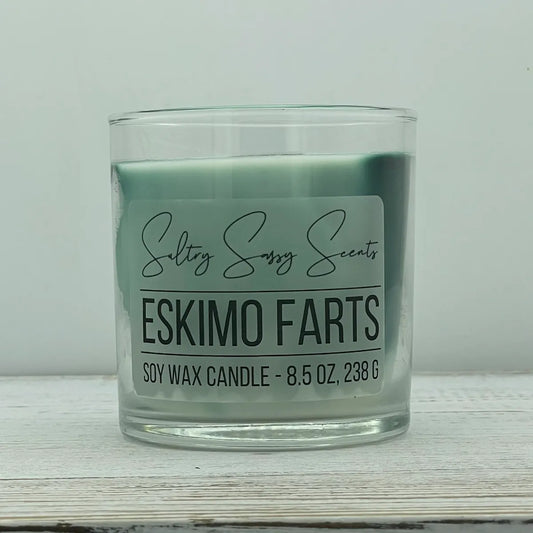 An 8.5-ounce clear glass jar of Sultry Sassy Scents "Eskimo Farts" soy wax candle featuring a smooth, pale teal-blue wax color and a flat wooden wick, centered on a white wood surface.