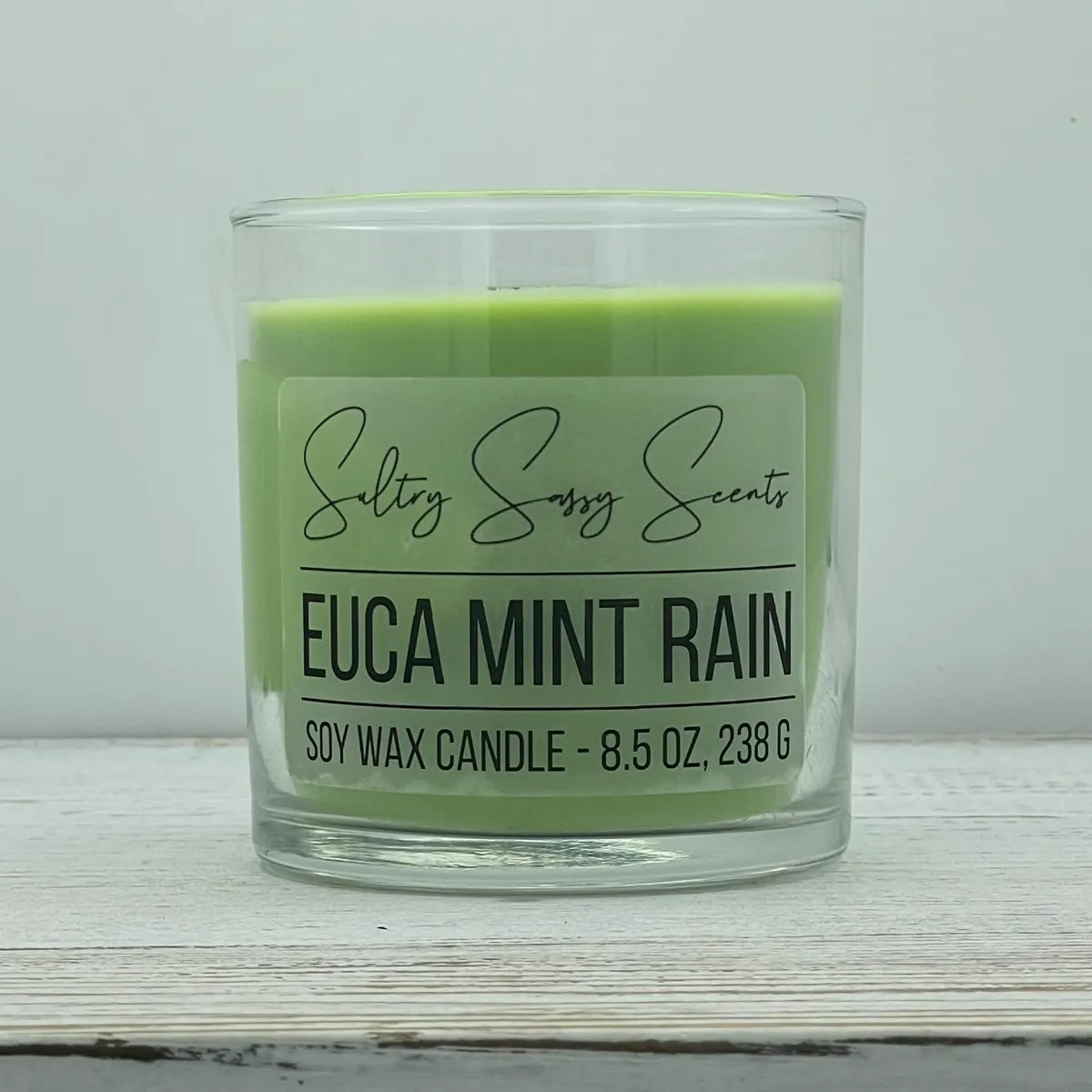 An 8.5-ounce clear glass jar of Sultry Sassy Scents "Euca Mint Rain" soy wax candle displaying a soft pastel sage green wax color, centered against a light gray background.