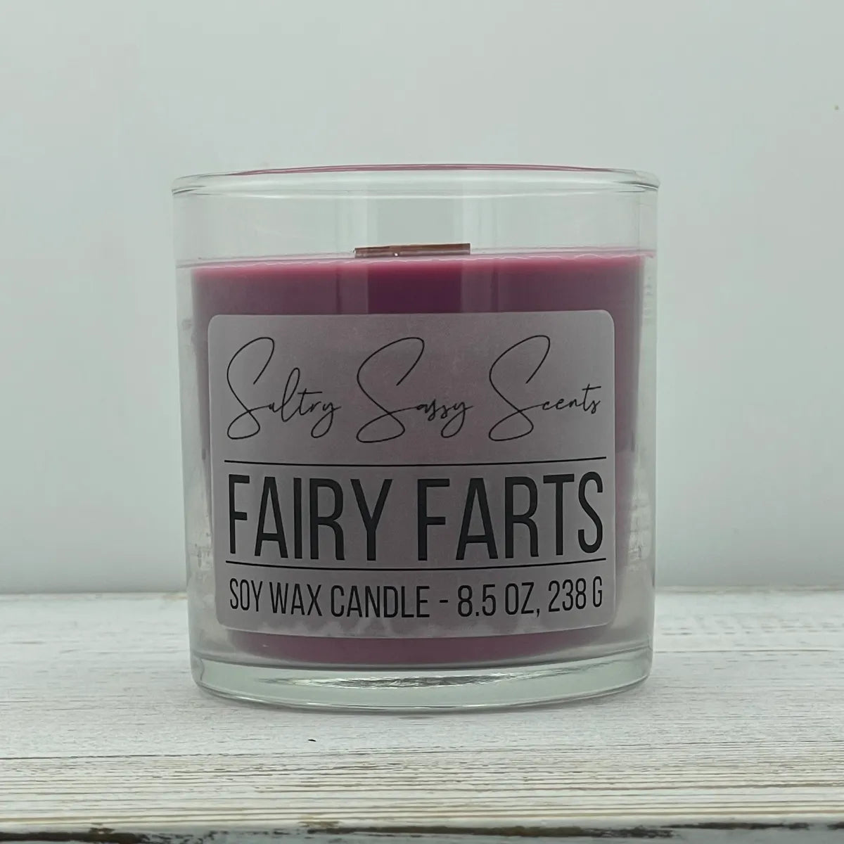 An 8.5-ounce clear glass jar of Sultry Sassy Scents "Fairy Farts" soy wax candle showcasing a deep mauve-purple wax color with a flat wooden wick, resting on a white-washed wood table.