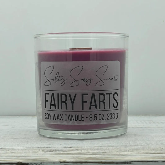 An 8.5-ounce clear glass jar of Sultry Sassy Scents "Fairy Farts" soy wax candle showcasing a deep mauve-purple wax color with a flat wooden wick, resting on a white-washed wood table.