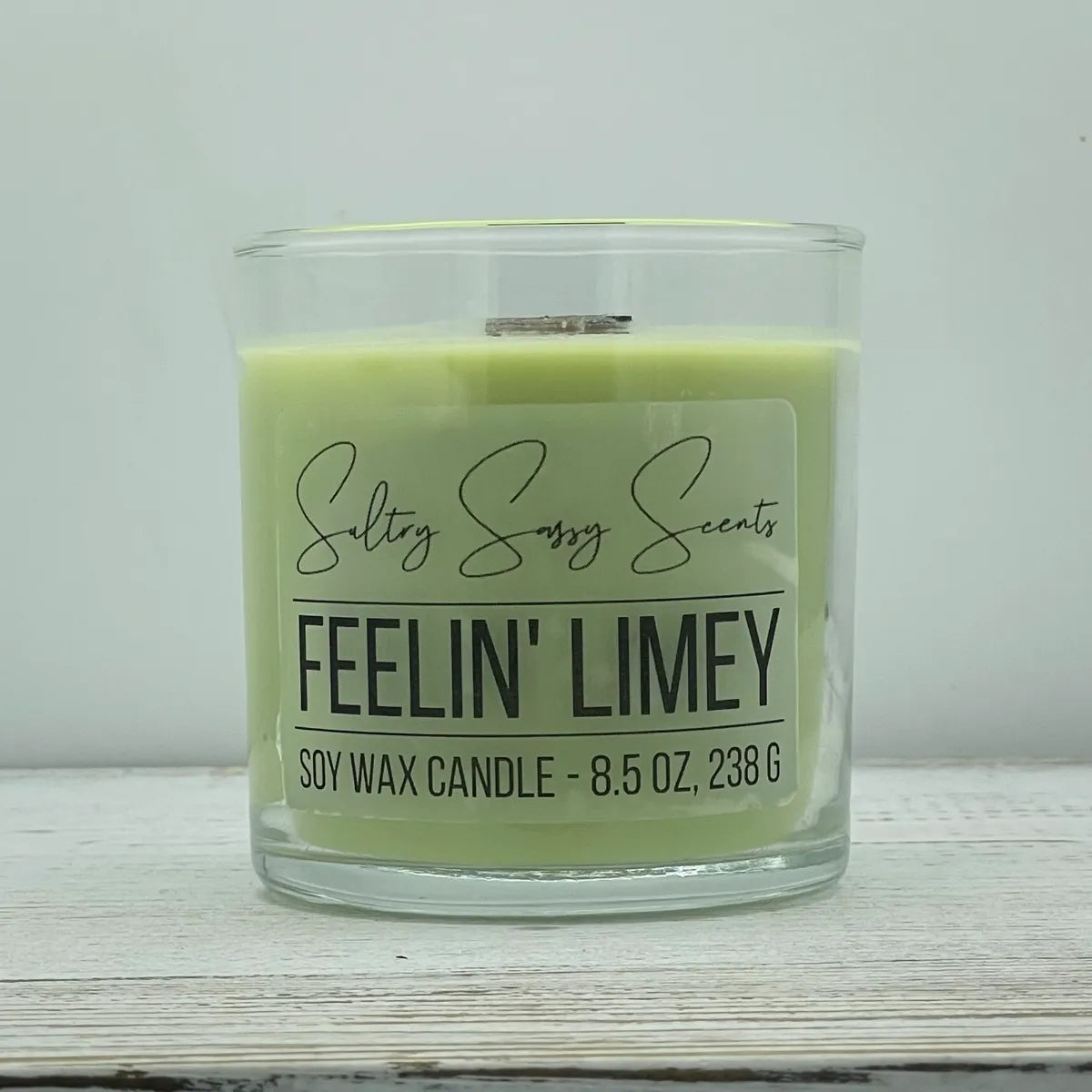An 8.5-ounce clear glass jar of Sultry Sassy Scents "Feelin' Limey" soy wax candle featuring a bright lime green wax color, displayed on a light wooden background.