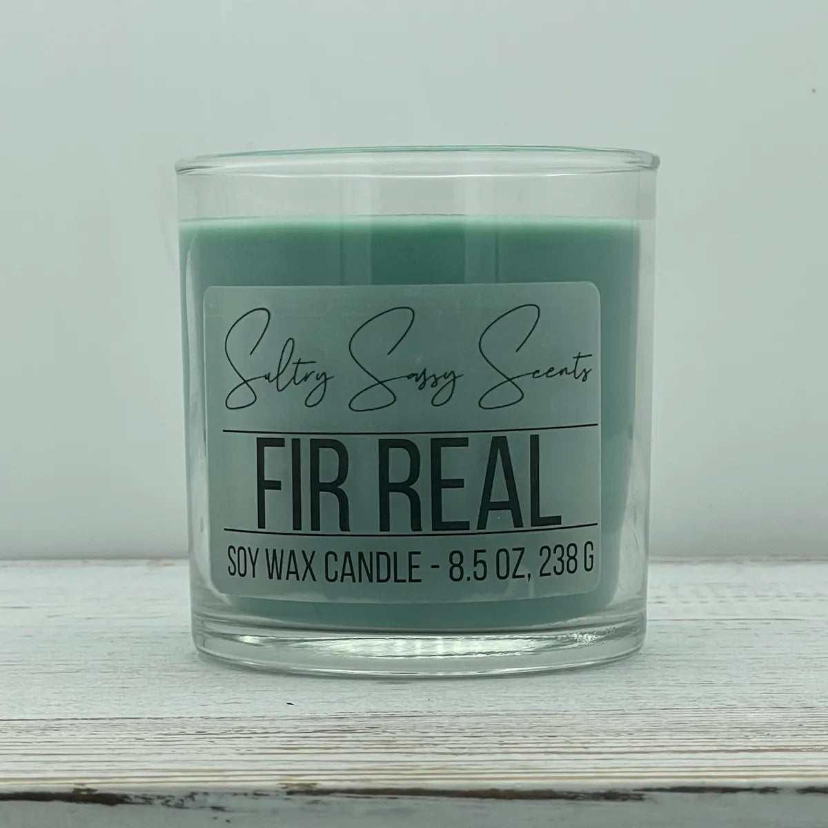 An 8.5-ounce clear glass jar of Sultry Sassy Scents "Fir Real" soy wax candle showcasing a deep muted pine green wax color with a flat wooden wick, positioned against a clean light background.