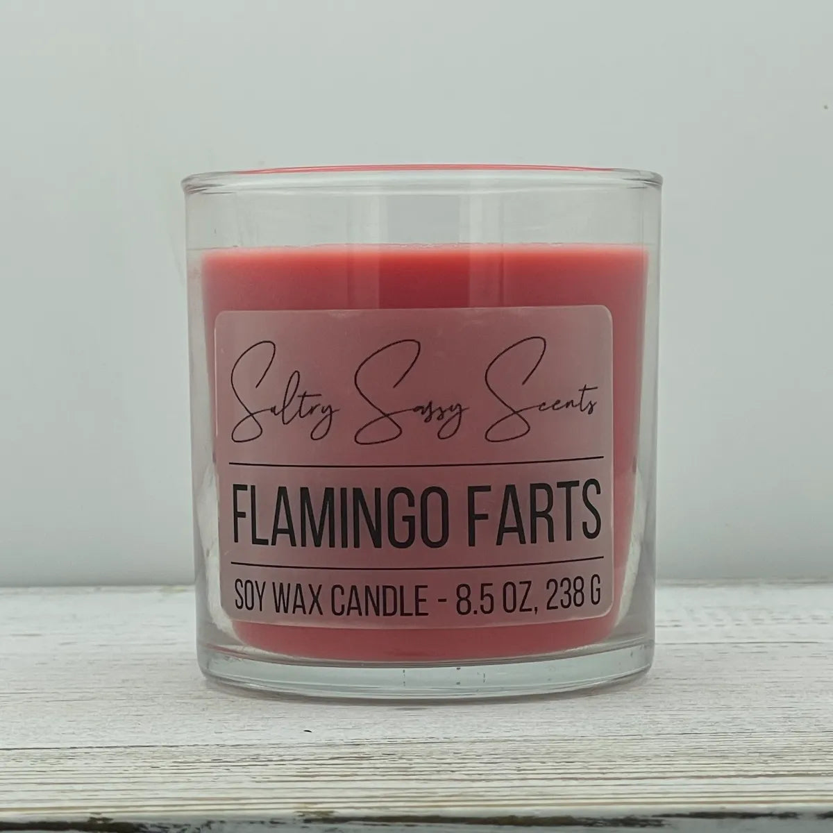 An 8.5-ounce clear glass jar of Sultry Sassy Scents "Flamingo Farts" soy wax candle highlighting a coral pink wax color, centered on a light wood surface.