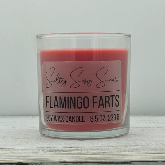 An 8.5-ounce clear glass jar of Sultry Sassy Scents "Flamingo Farts" soy wax candle highlighting a coral pink wax color, centered on a light wood surface.