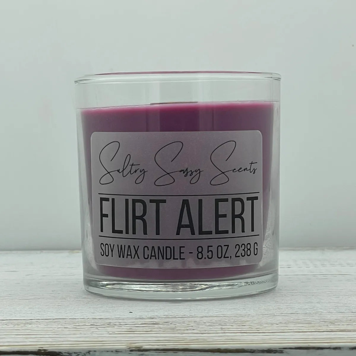 An 8.5-ounce clear glass jar of Sultry Sassy Scents "Flirt Alert" soy wax candle featuring a deep plum-purple wax color and a flat wooden wick.