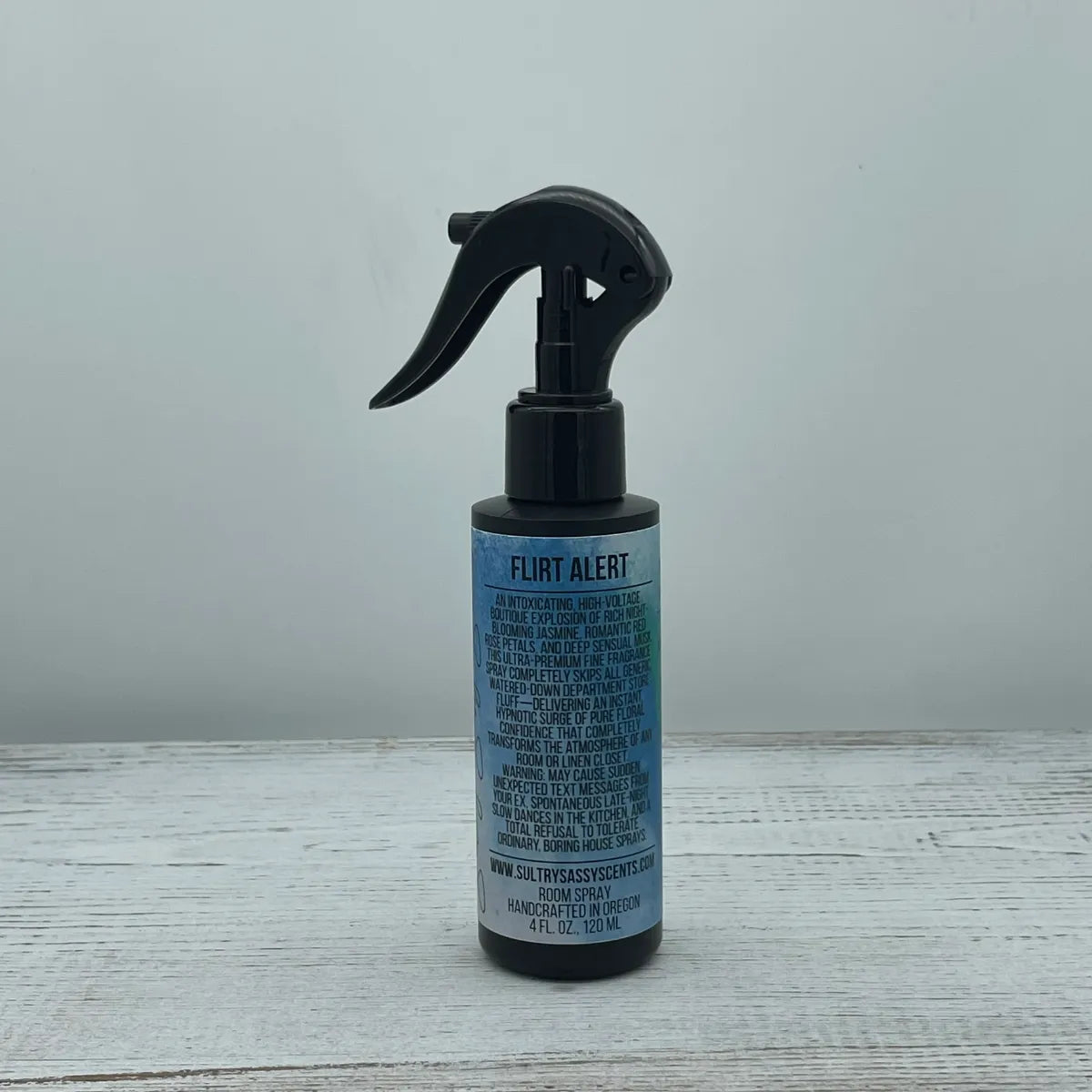 Front label view of a 4-ounce black spray bottle for Sultry Sassy Scents "Flirt Alert" Room Spray, showcasing a full paragraph description with fragrance notes on a white wooden table.