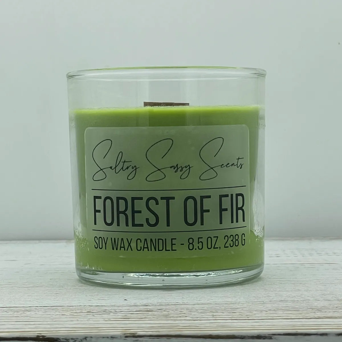 An 8.5-ounce clear glass jar of Sultry Sassy Scents "Forest of Fir" soy wax candle featuring a rich pine green wax color and a flat wooden wick, centered on a white wood surface.