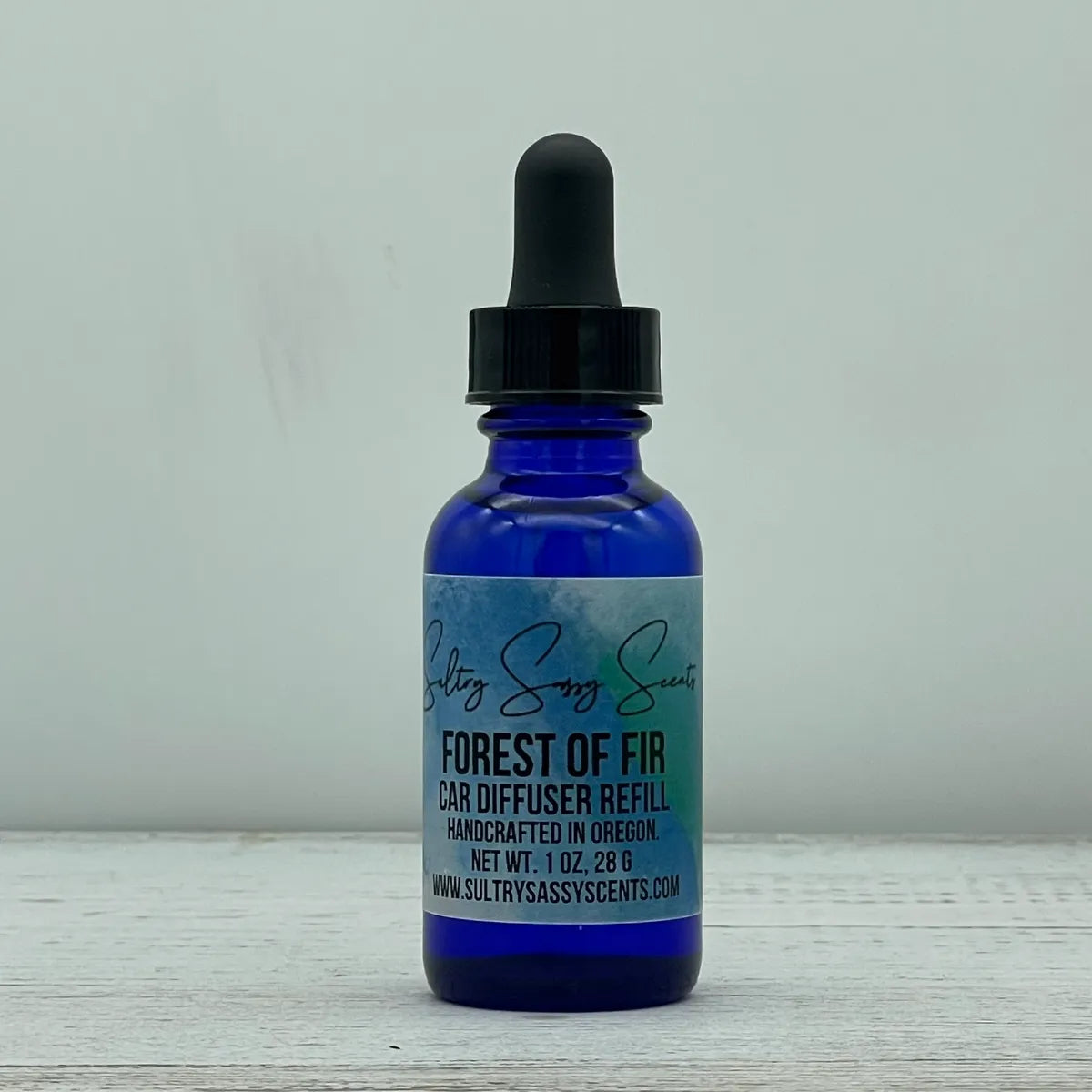 Front label view of a cobalt blue glass dropper bottle containing Sultry Sassy Scents "Forest of Fir" Car Diffuser Refill, resting on a white-washed wood grain surface.