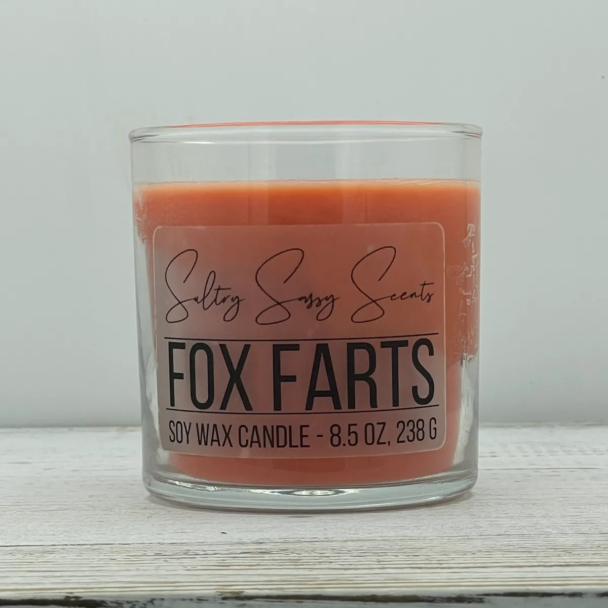An 8.5-ounce clear glass jar of Sultry Sassy Scents "Fox Farts" soy wax candle showcasing a smooth rust orange wax color and a flat wooden wick.